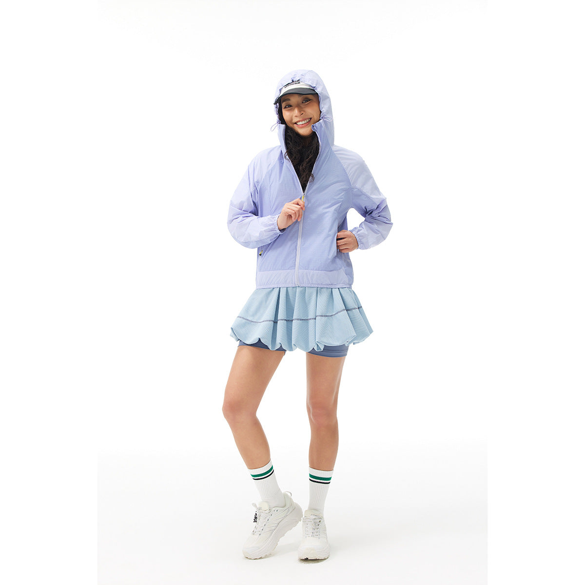 Woman wearing a lavender hooded zip jacket with a pale blue ruffled athletic skirt and shorts.