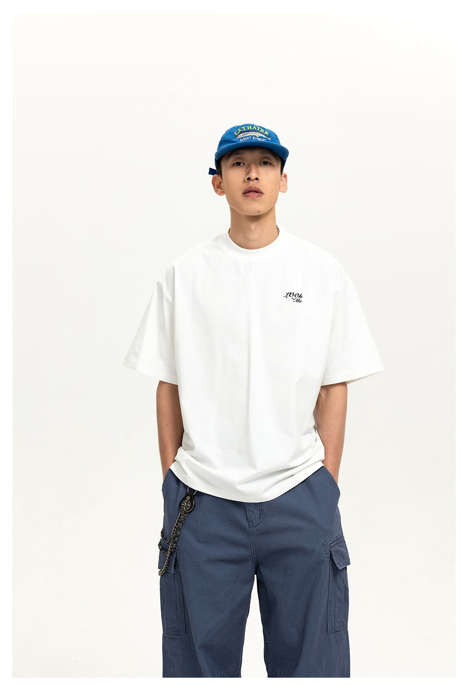 Model wearing a white short-sleeve T-shirt with small chest script, blue cap and blue cargo pants