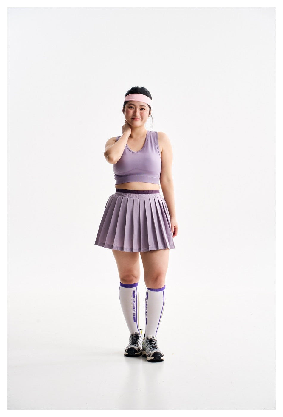 Full front view of a model wearing a lavender sleeveless sports bra, pleated skirt, pink headband, knee-high socks, and