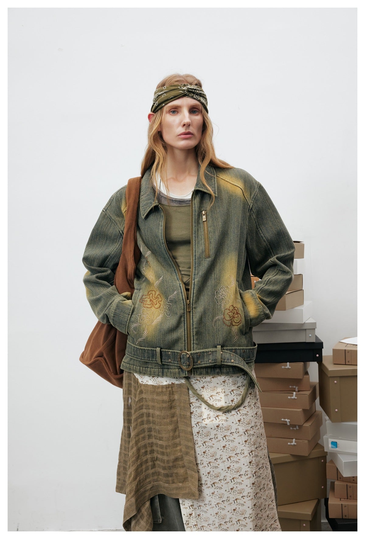 Model wearing a distressed olive jacket with floral embroidery, zip closure, and belt detail at the hem.
