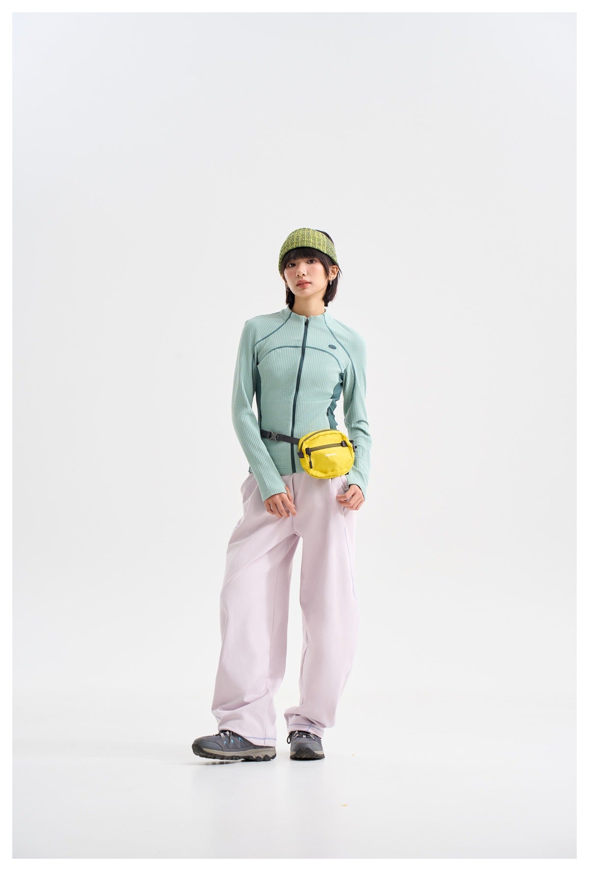 Model wearing a pale aqua zip-front jacket, loose lavender pants, and a bright yellow waist pack.