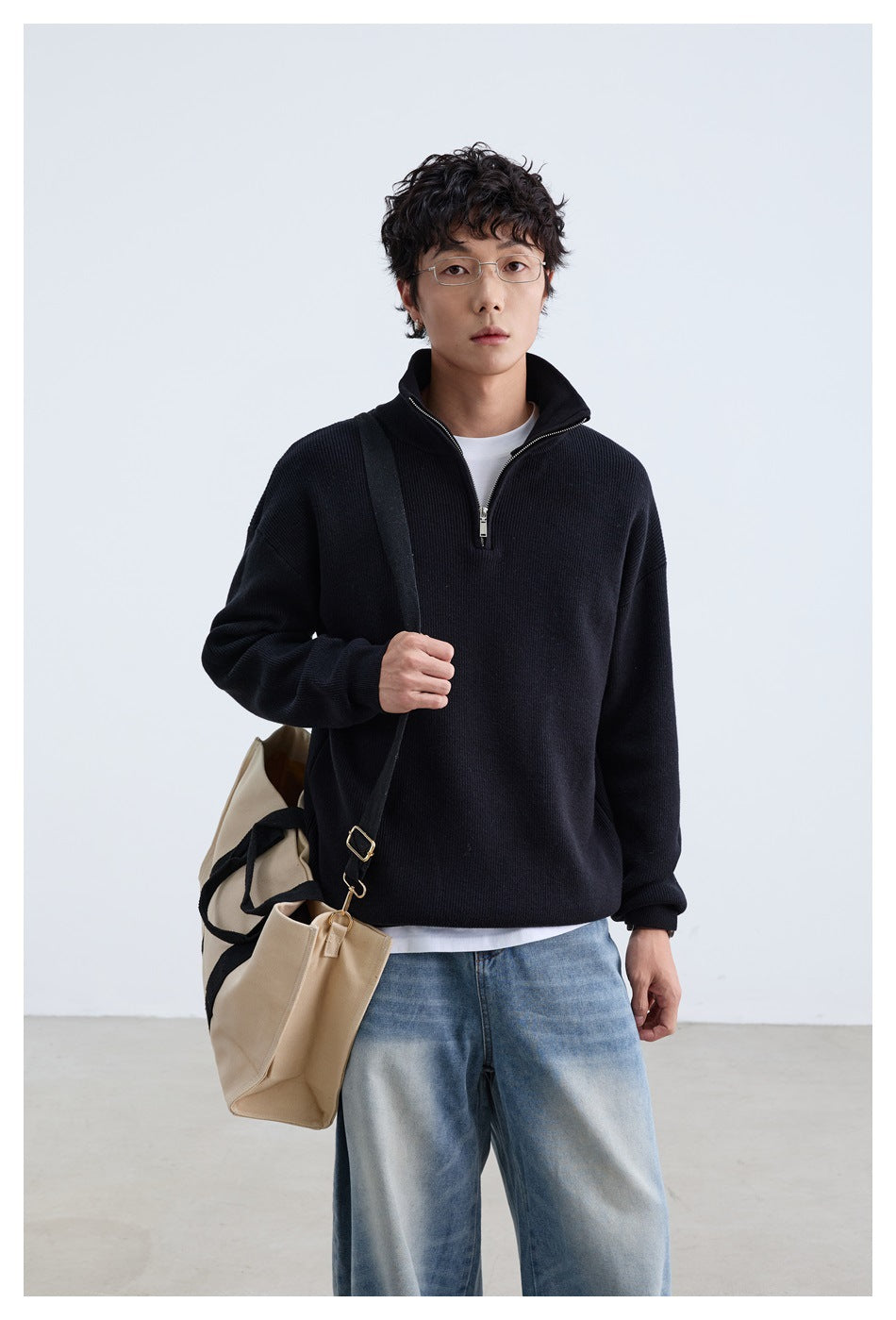 Front view of a model in a black quarter-zip sweater and blue jeans, carrying a beige tote.