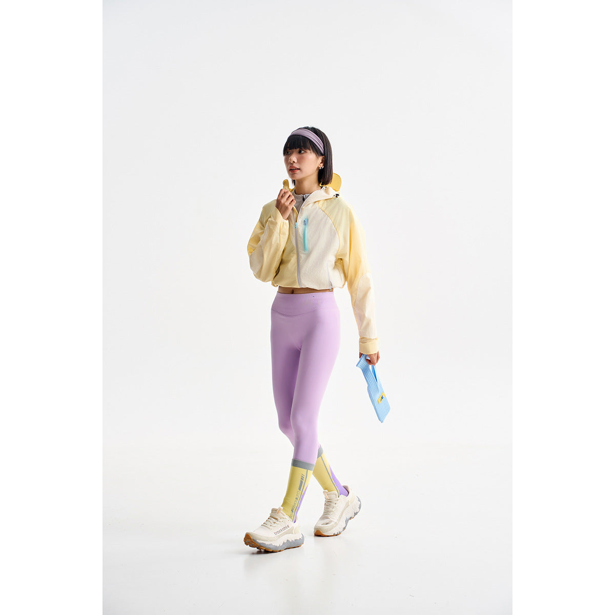 Model wearing a pale yellow cropped hooded jacket with lavender leggings and yellow-accented socks.
