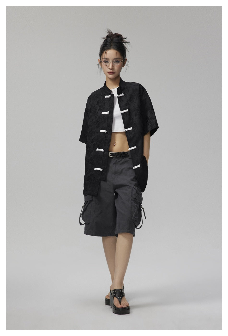 Full-length view of a model wearing a black short-sleeve jacket with white frog closures and dark cargo shorts.