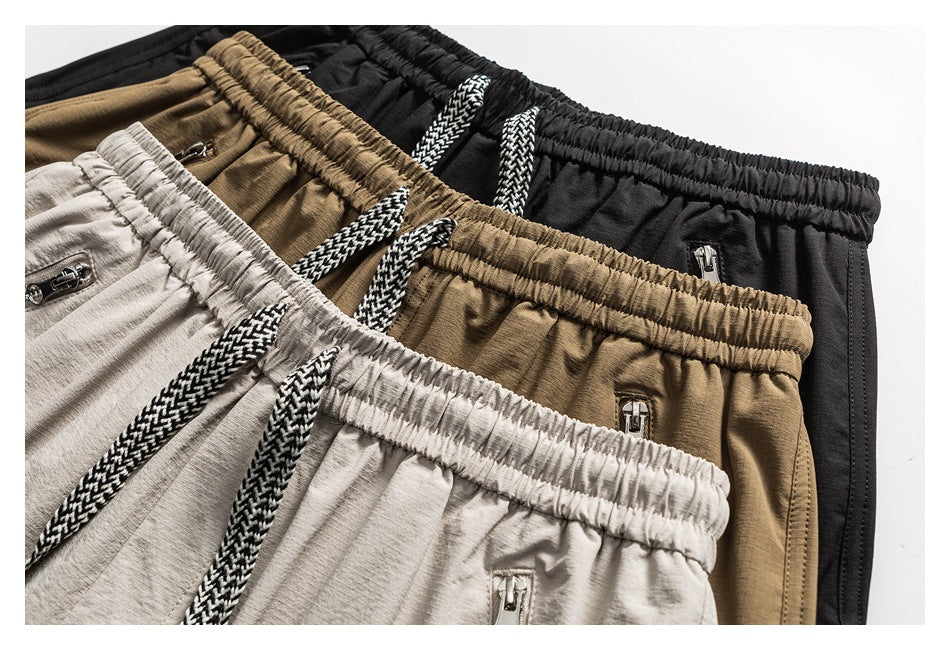 Three pairs of shorts in black, tan, and light gray, showing elastic waistbands, drawstrings, and zip pockets.
