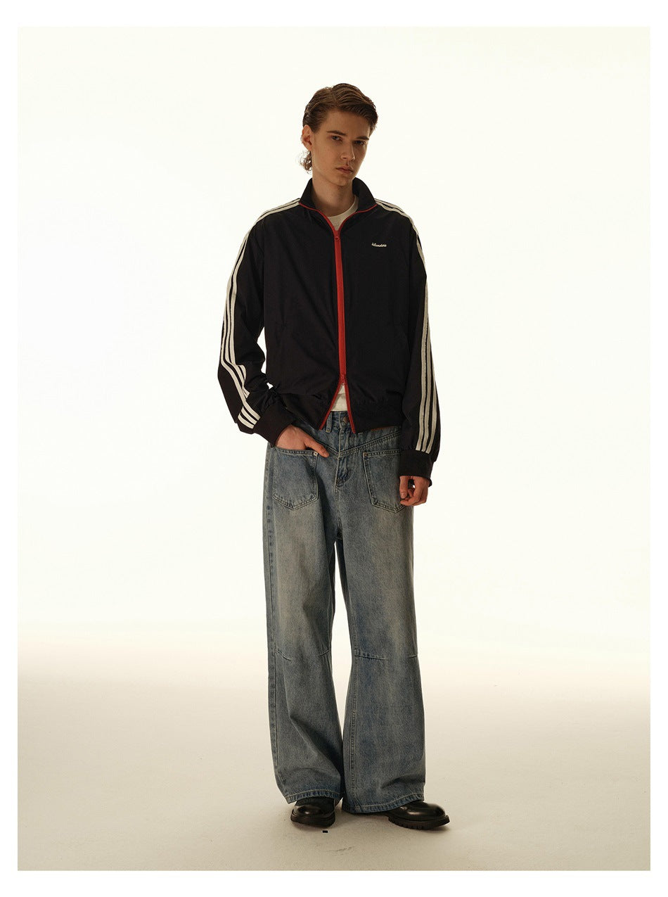 Man wearing a black track jacket with white sleeve stripes and red zipper trim, paired with loose gray jeans.