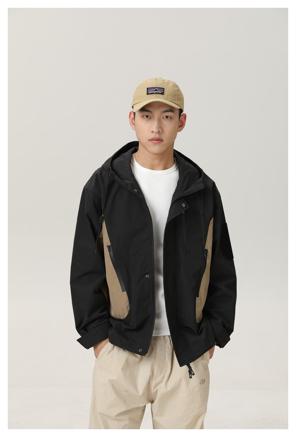 Man in a black hooded jacket with khaki side panels worn open over a white tee, front view