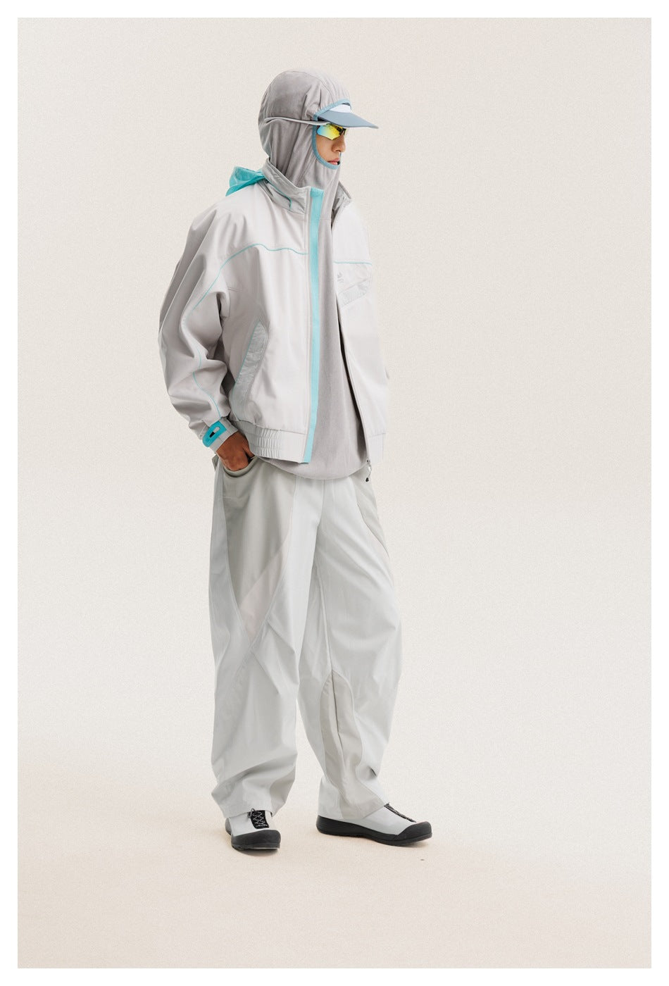 Side view of a model in a light gray jacket with turquoise trim and loose matching pants, wearing a hood and cap.
