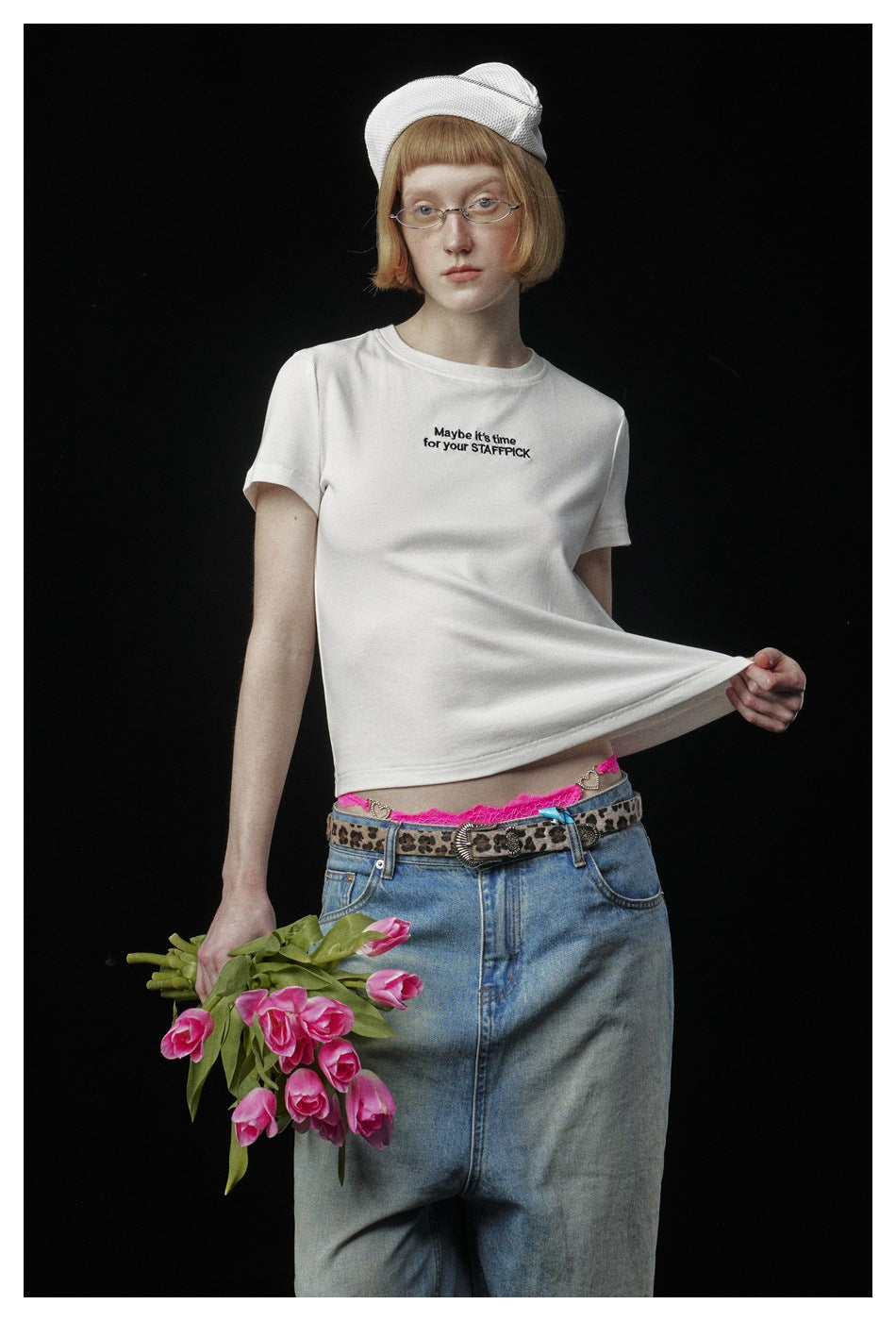 Model wearing a white short-sleeve T-shirt with small black chest text, paired with a denim skirt and white cap.