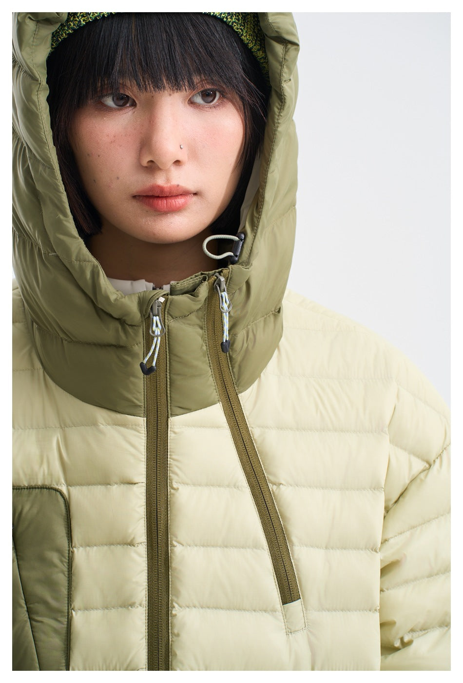 Close-up of a model wearing a cream quilted jacket with an olive hood, drawcords and contrasting front zippers.