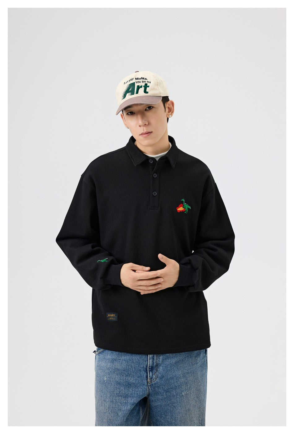 Model wearing a black collared pullover with colorful embroidered motifs on the chest and sleeve, hands clasped.