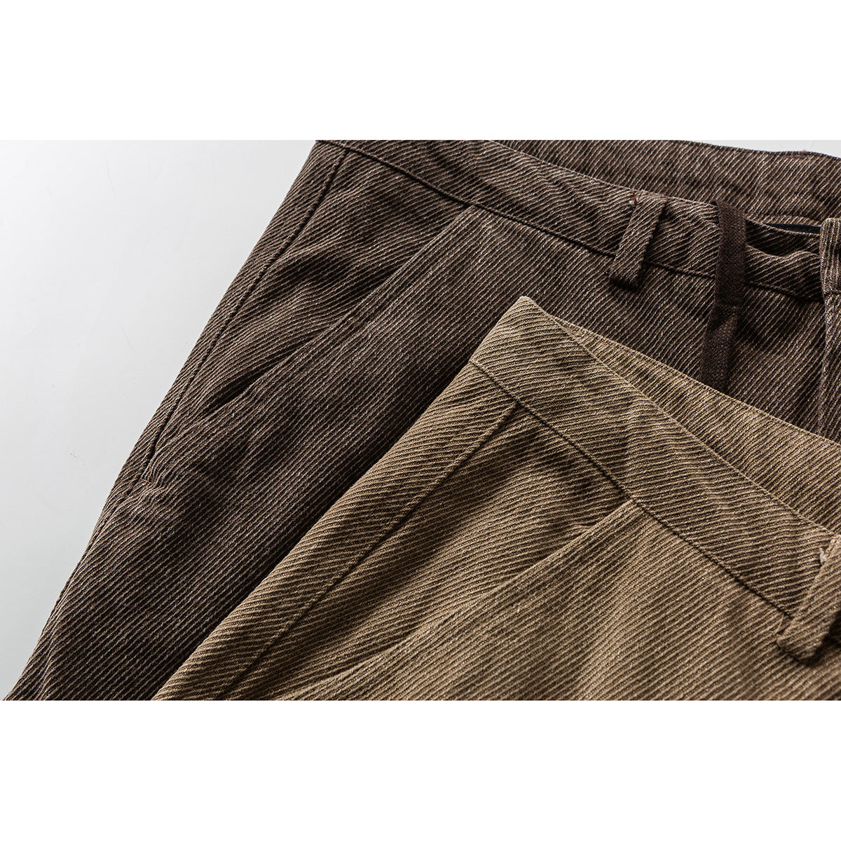 Close-up of two brown and tan trousers showing diagonal fabric texture, waistbands and belt loops.
