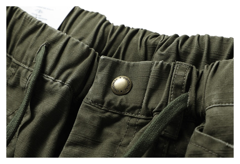 Close-up of the gathered waistband, drawstring and metal button stamped “ORIGINAL” on olive-green pants.