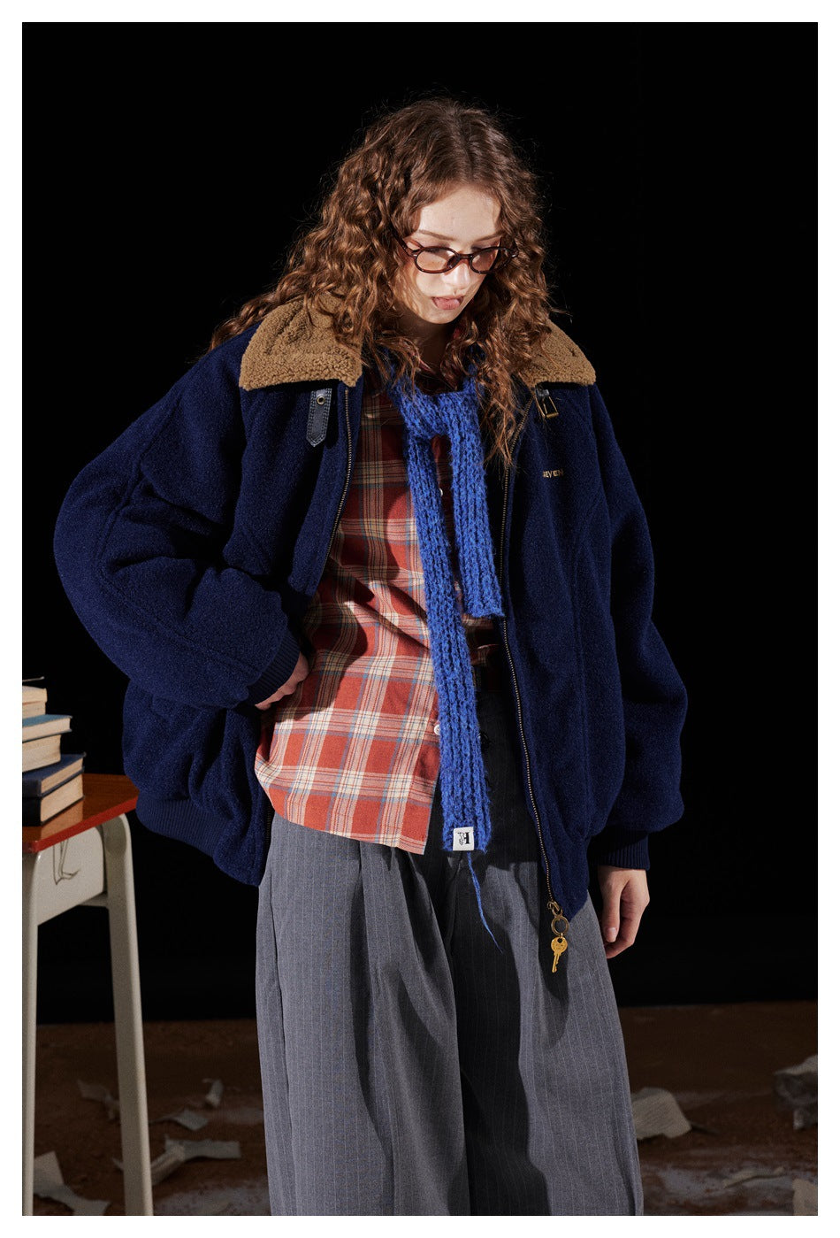Model wearing a navy zip jacket with tan fleece-like collar over a plaid shirt, blue scarf and gray striped trousers