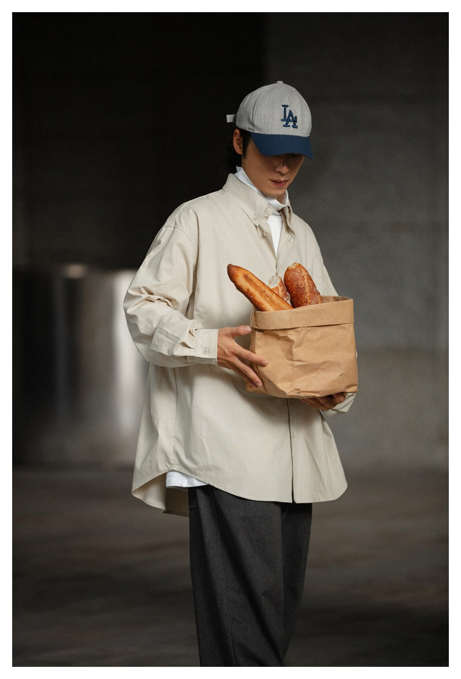 Model wearing a loose cream button-up shirt and dark trousers, holding a paper bag of bread.