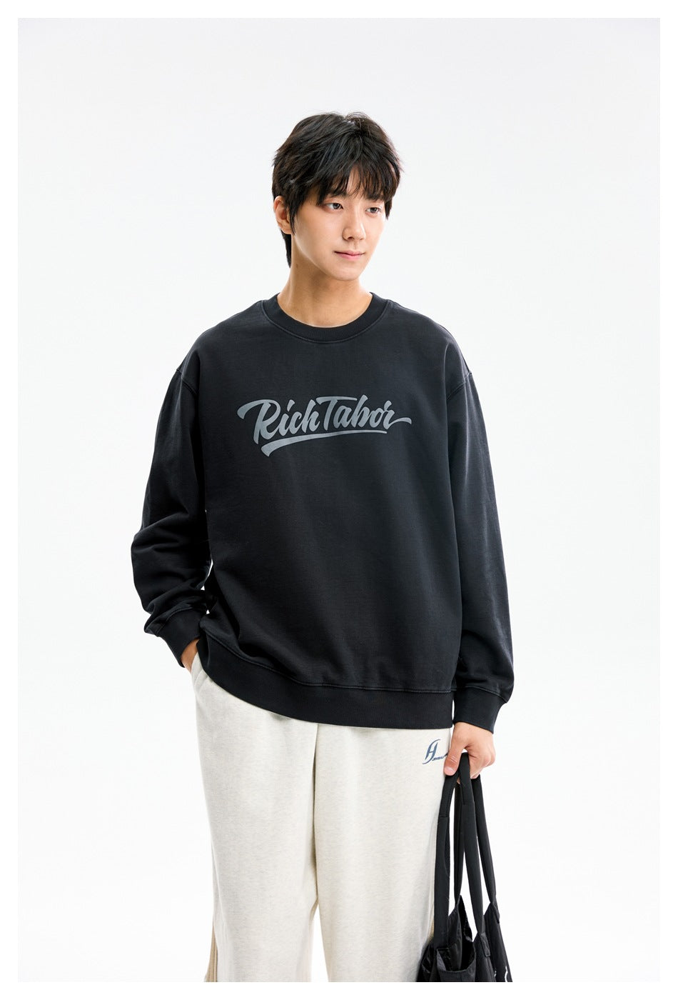 Model wearing a black crewneck sweatshirt with a large gray script print across the chest.