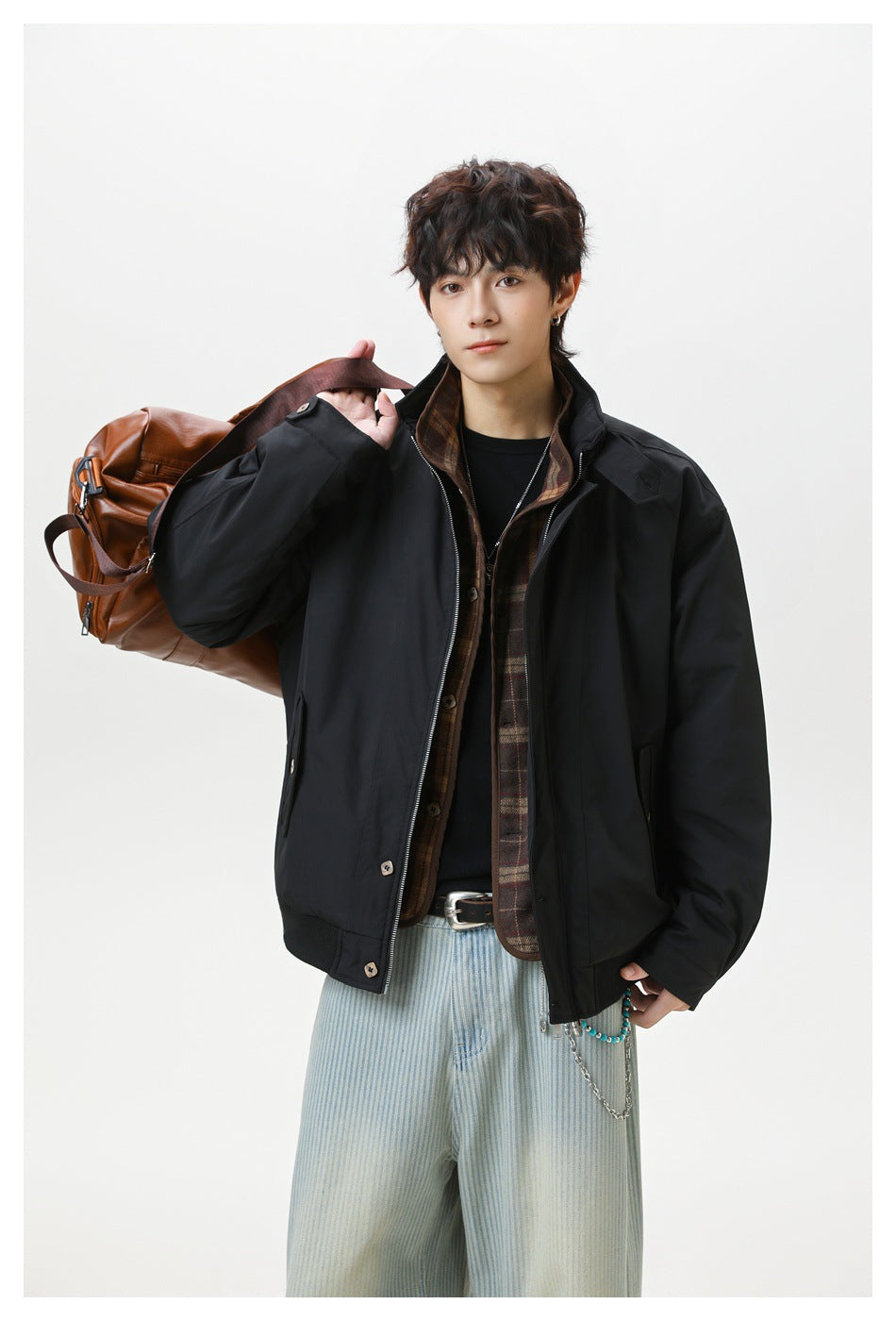 Model wearing a black zip-front jacket over a brown plaid layer, carrying a brown leather-look bag over one shoulder.
