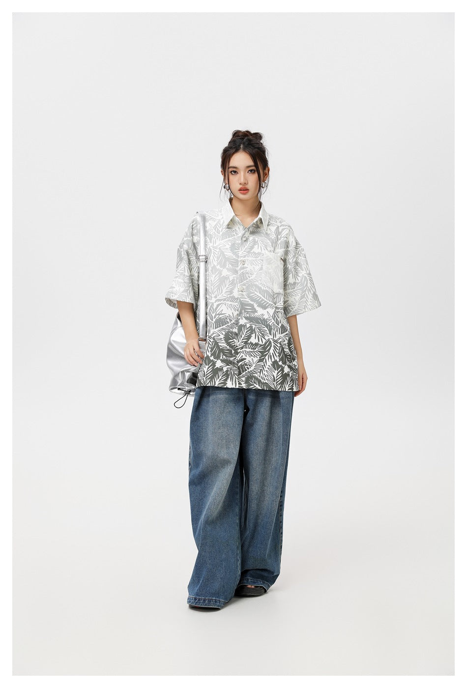 Front view of a model wearing a gray-and-white tropical leaf print short-sleeve shirt with wide blue jeans and a silver bag.