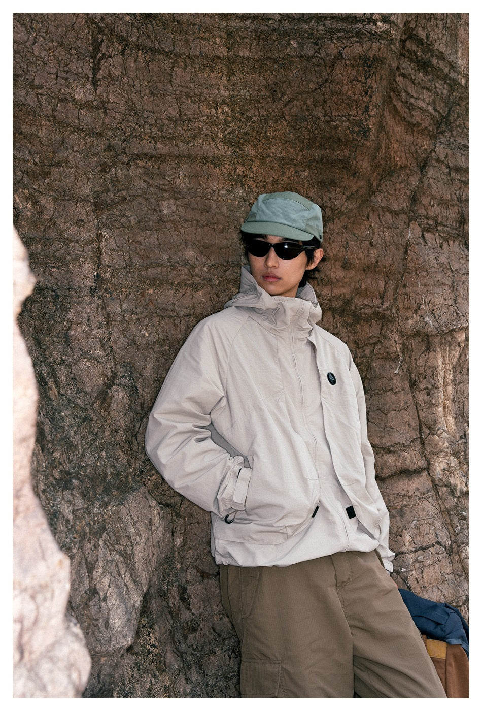 A model poses against a rock wall wearing a light beige hooded jacket, olive trousers, and a sage cap.