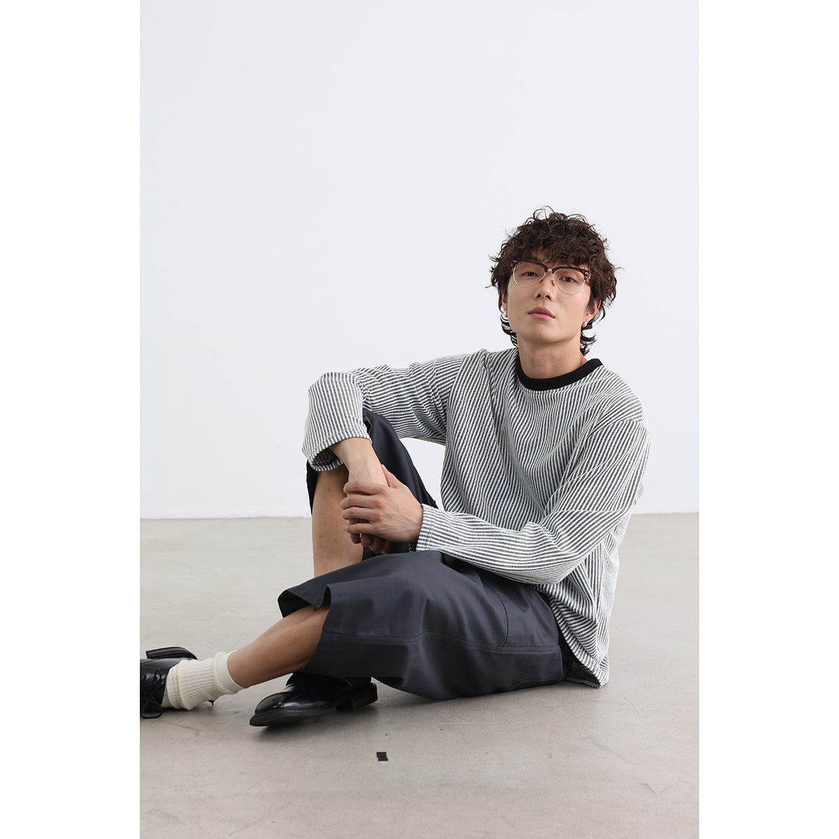 Seated model wearing a gray-and-white striped long-sleeve crewneck with dark shorts.