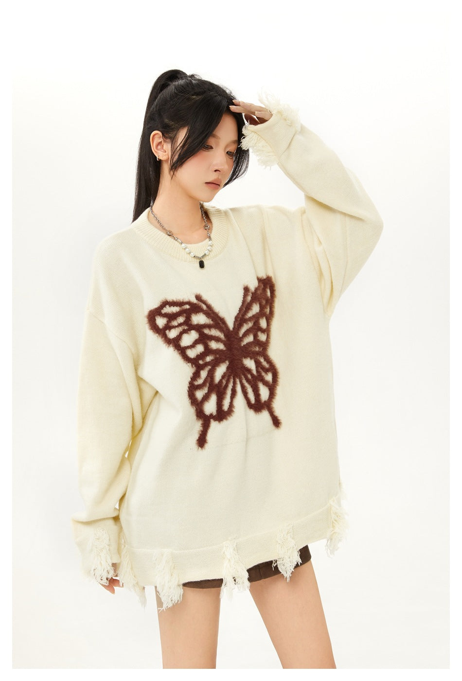 Angled view of a model in an oversized ivory sweater with a brown butterfly motif and fringe at the cuffs and hem.