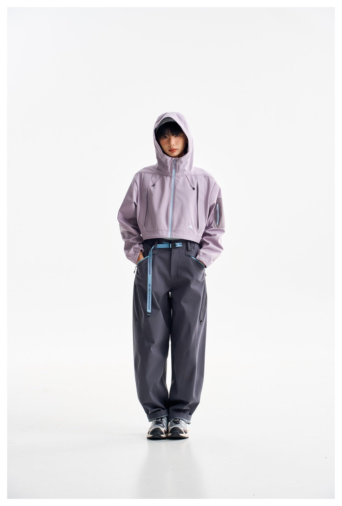 Model wearing a lavender cropped hooded jacket with the hood up and dark loose trousers against a white background.