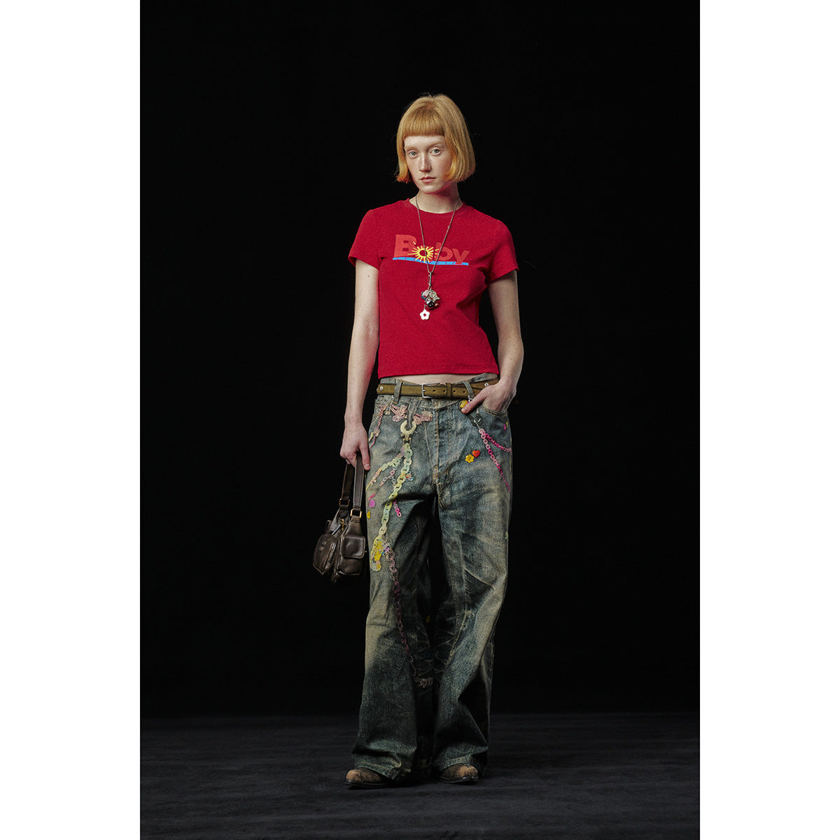 Model wearing a red graphic T-shirt with loose decorated jeans, photographed against a black background.