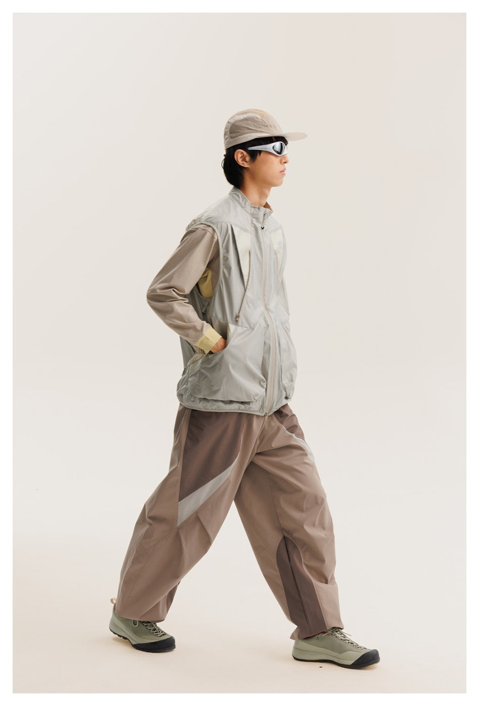 Side view of a model in a light gray sleeveless zip vest, taupe pants, cap and sport sunglasses.