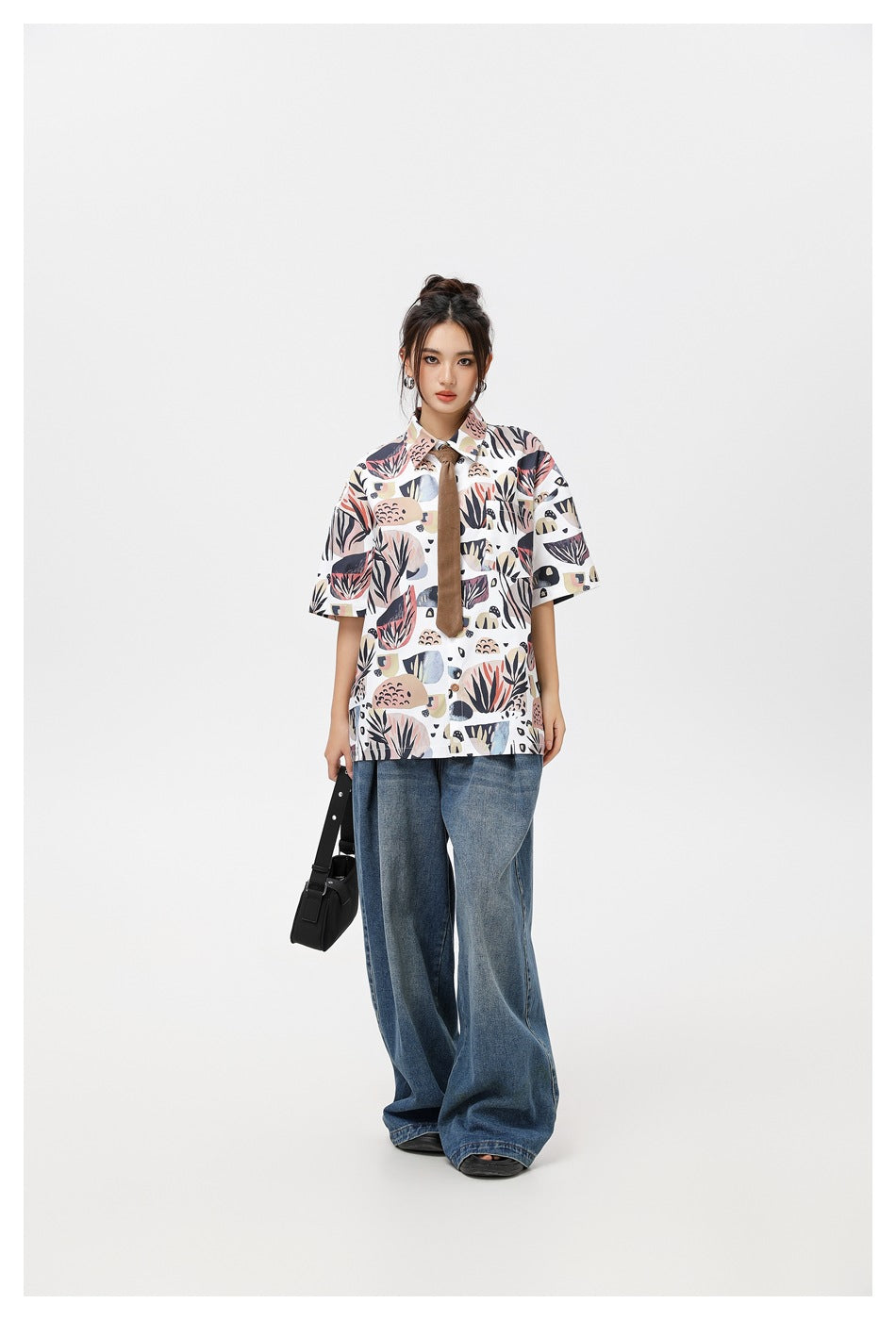 Full-length front view of a model in a white botanical-print short-sleeve shirt, brown tie, and wide-leg blue jeans.