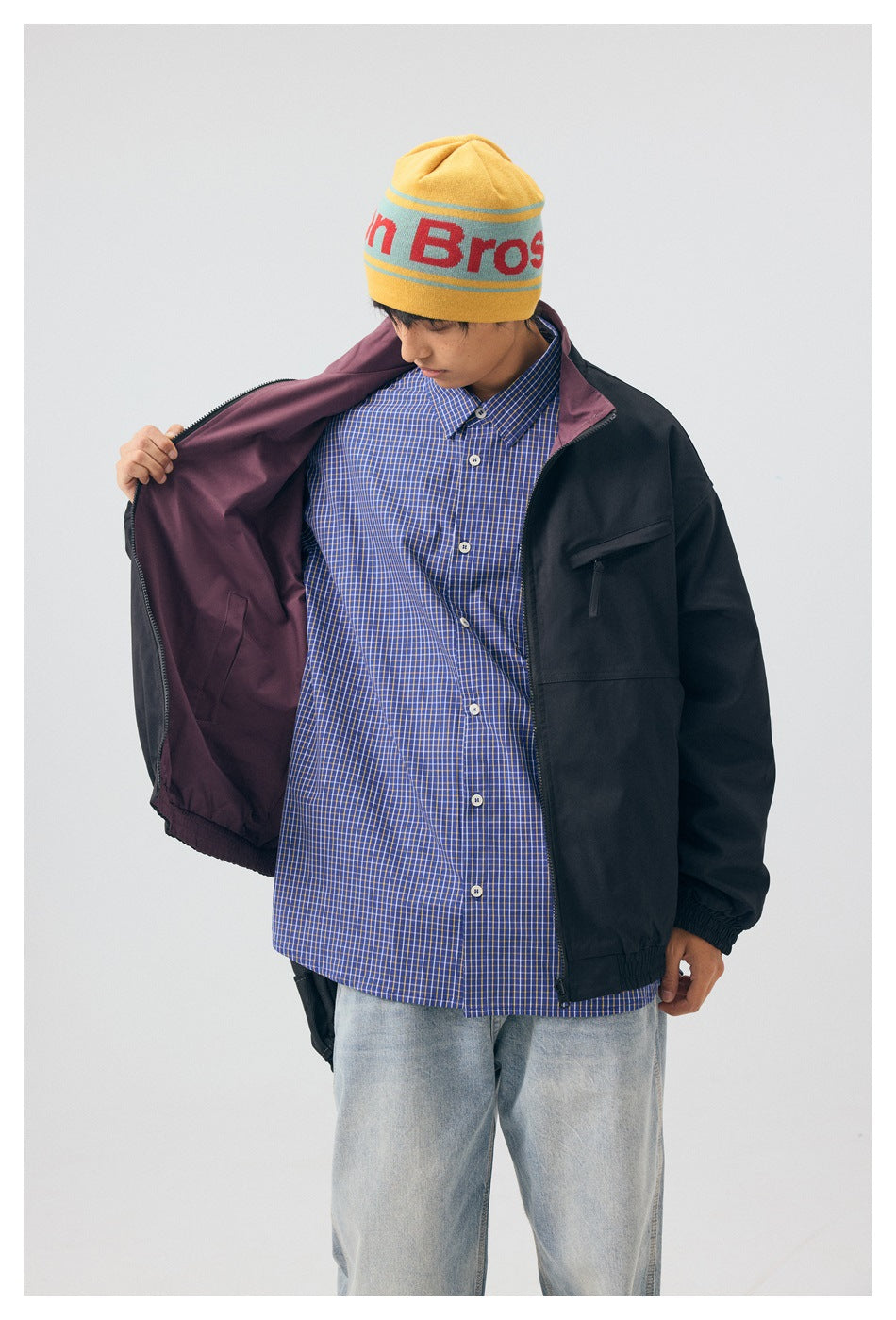 Model holds open a black jacket to show its plum lining, over a blue checked shirt.