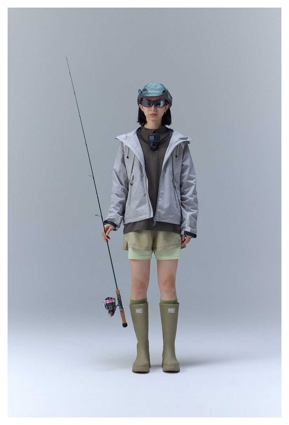 Model wearing the light gray hooded jacket open over a dark top with shorts, boots and a fishing rod, front view