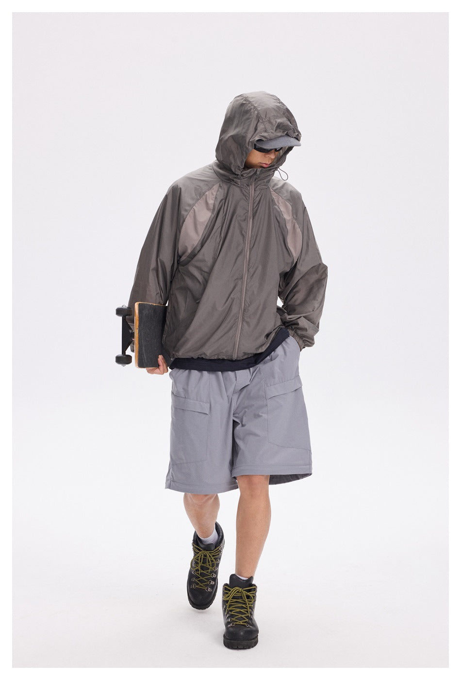 Model wearing a dark gray hooded jacket and gray shorts while carrying a skateboard.