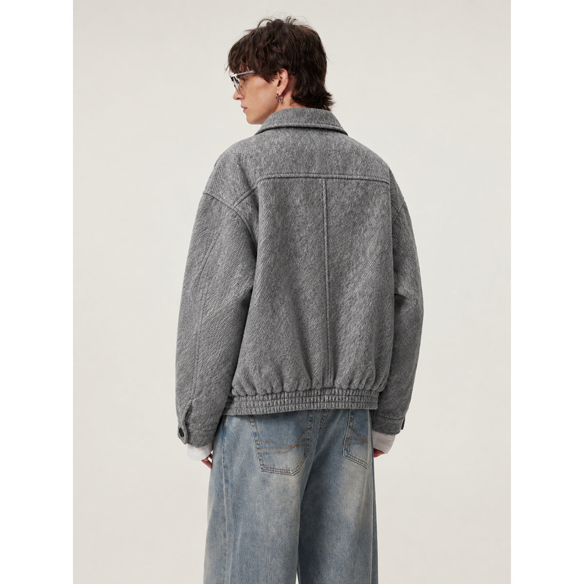 Rear view of a model wearing a gray textured jacket with a wide collar and gathered hem, paired with loose blue jeans.