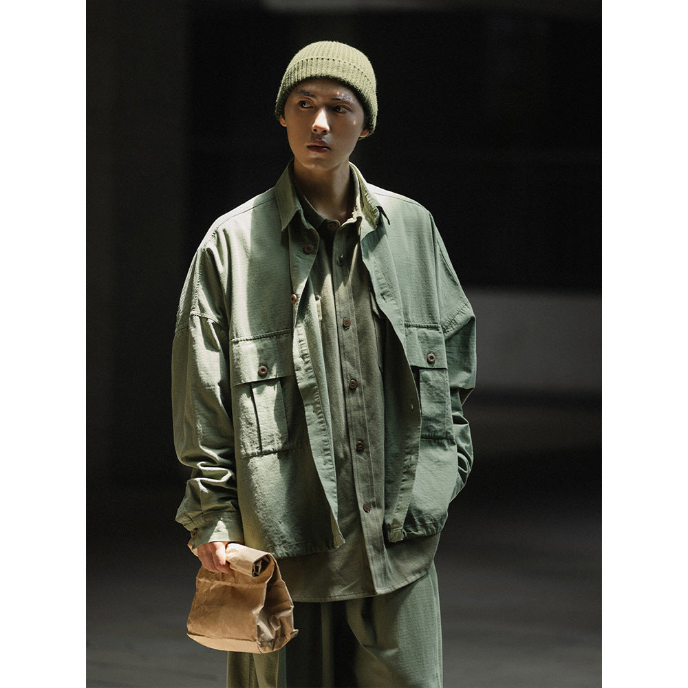 Model wearing a sage green oversized button-up jacket with flap pockets over matching layers and a cream knit beanie.