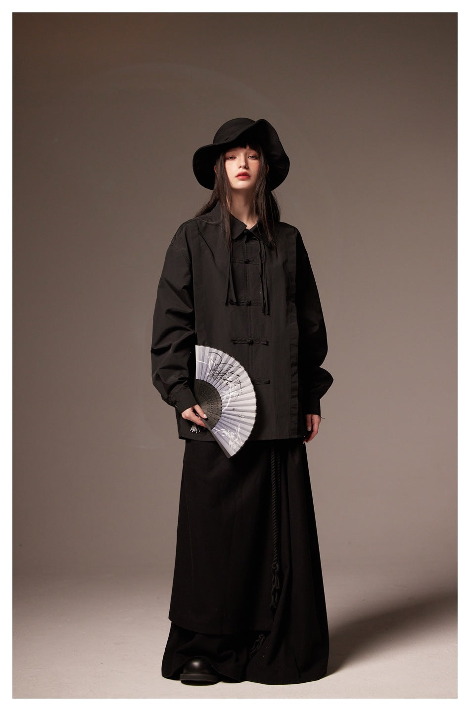 Full-length front view of a model in a black collared shirt with frog closures and a wide-brim hat.