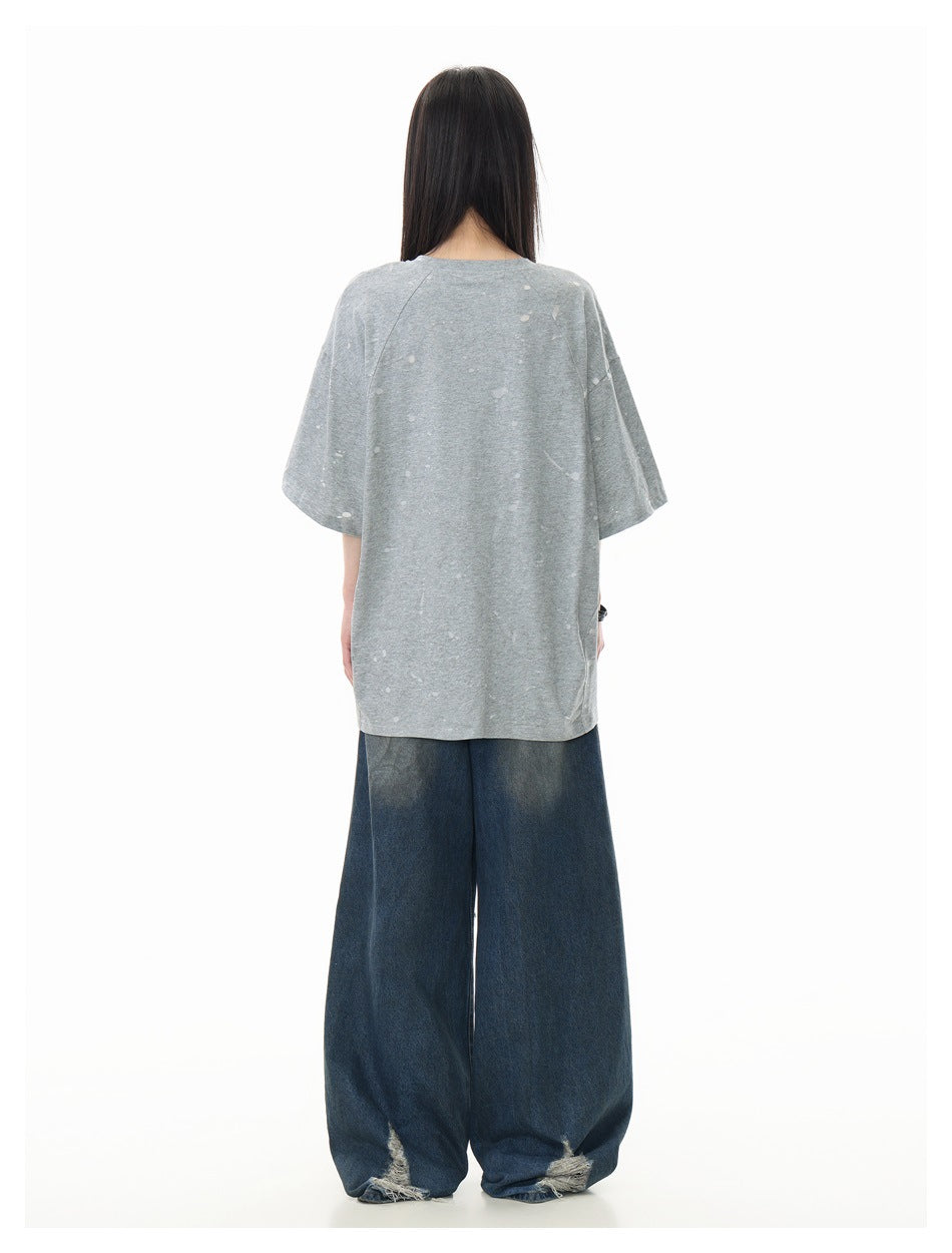 Back view of a model wearing a loose light-gray T-shirt with scattered pale marks and wide dark jeans.