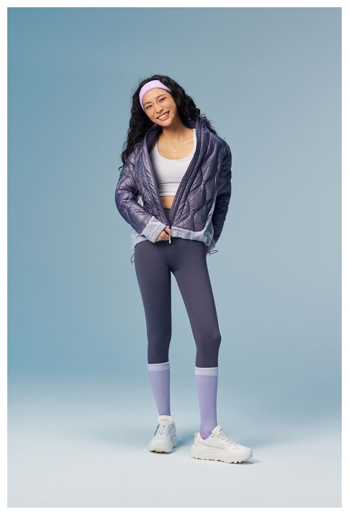Full-length model wearing an open purple quilted jacket, dark leggings, lavender socks, and white sneakers.