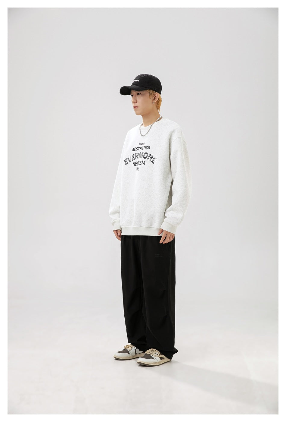 Man in light gray crewneck sweatshirt with front lettering print, black pants and cap, shown in three-quarter side view