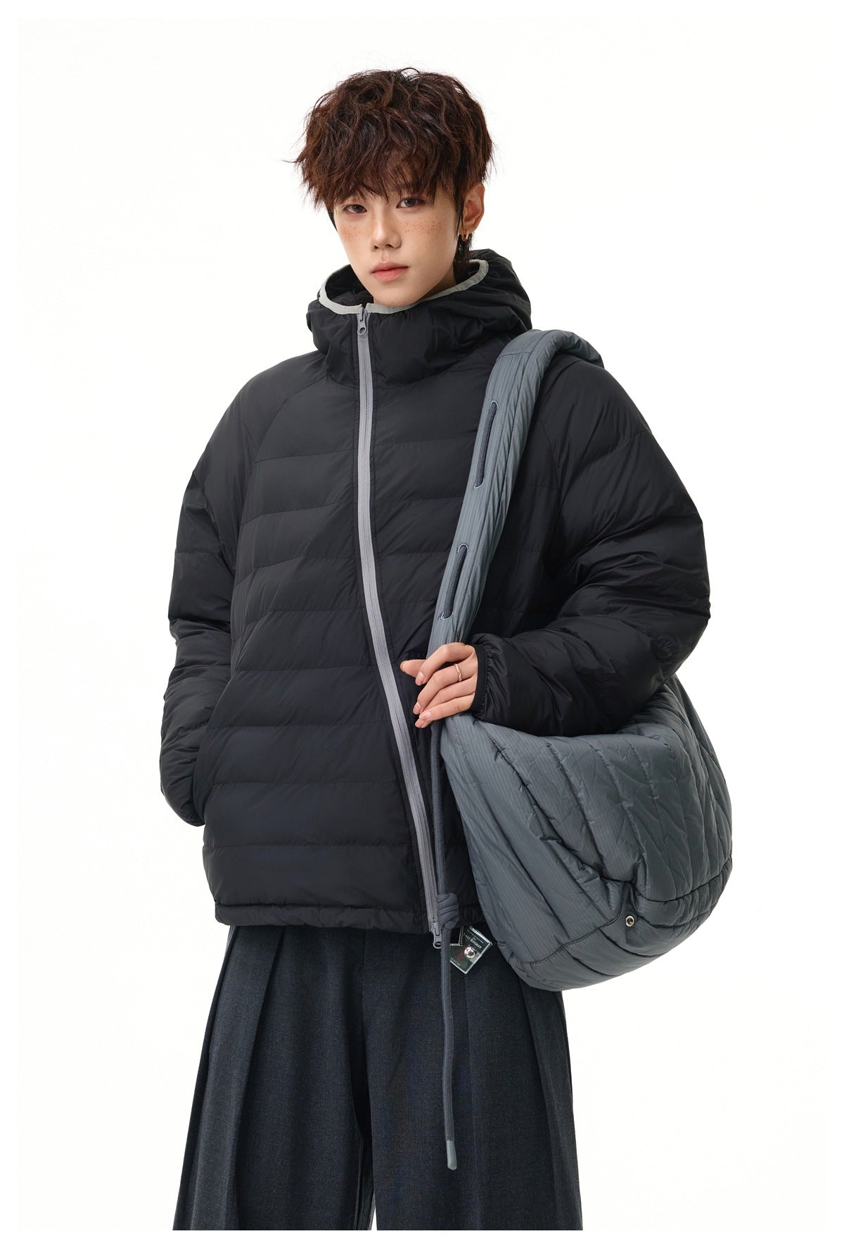 Model wearing a black hooded quilted puffer jacket and carrying a large gray quilted shoulder bag.