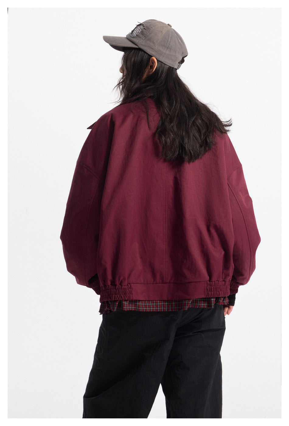 Rear view of a model wearing a burgundy jacket with gathered cuffs and hem, black trousers, and a gray cap.