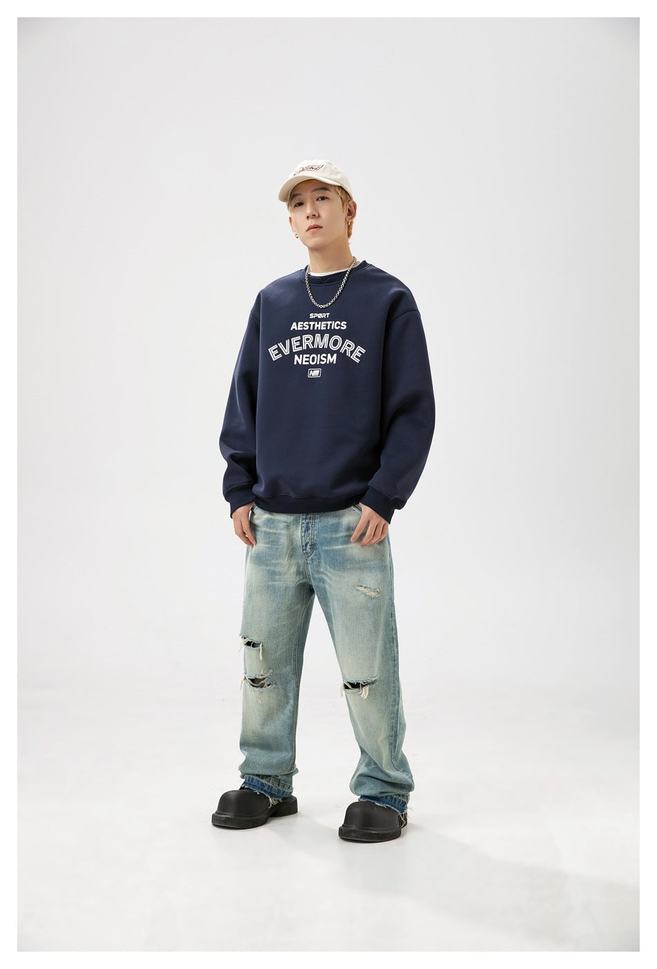 Model wearing a navy crewneck sweatshirt with white chest lettering, loose distressed jeans and a cream cap.