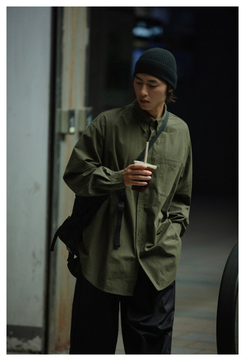 Model wearing an oversized olive-green button-up shirt with a chest pocket and black trousers.