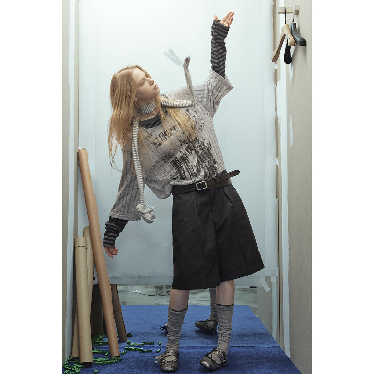 Model wearing a gray graphic top over striped sleeves with black knee-length shorts and a long striped scarf.