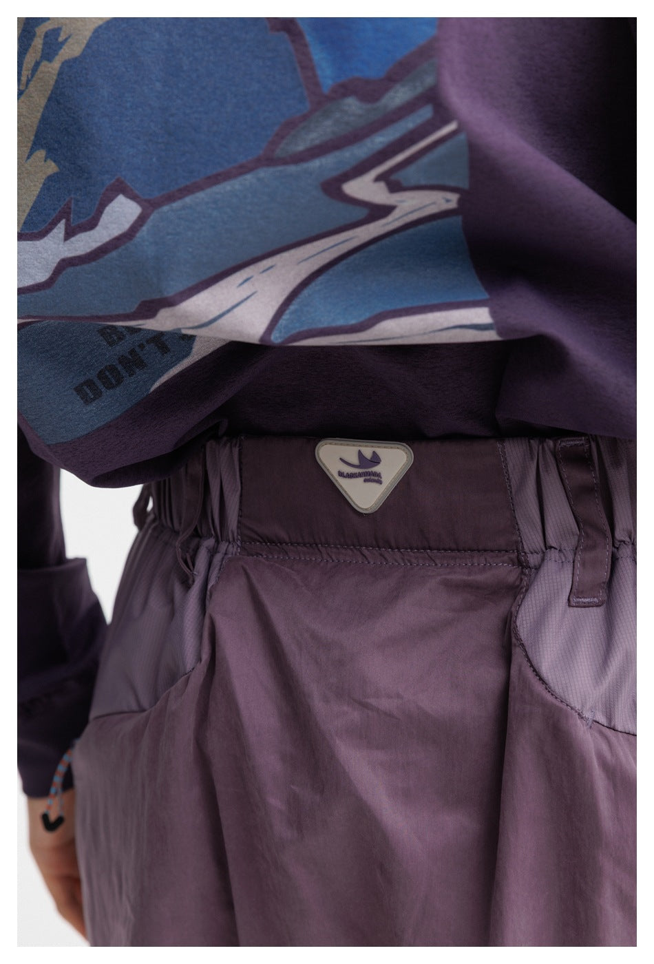 Close-up of the plum-purple pants' gathered waistband and branded badge, beside a colorful graphic top.