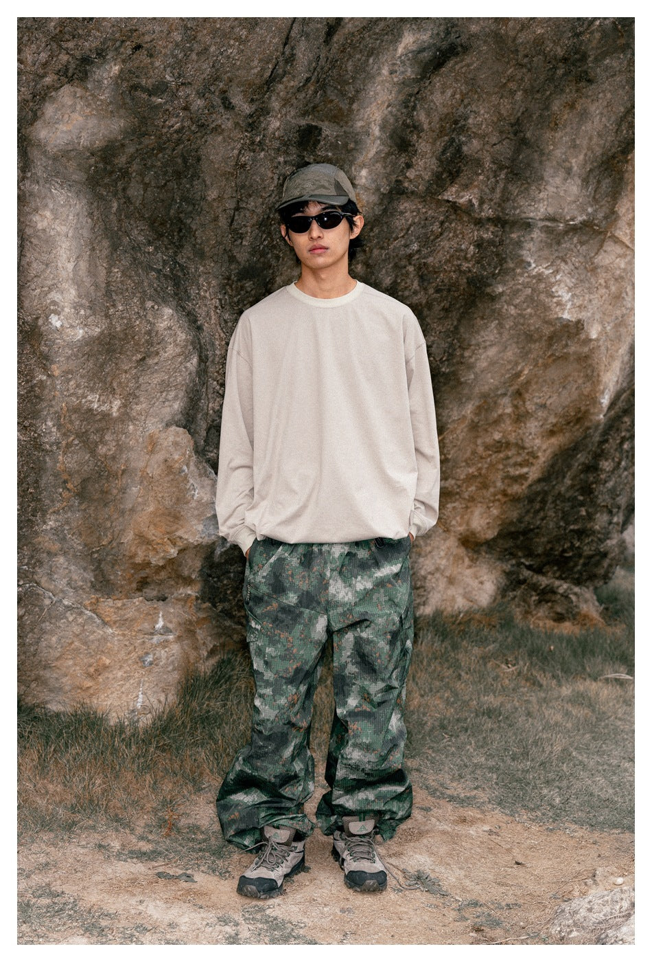Model facing forward in an oversized beige long-sleeve shirt and loose green camouflage trousers outdoors.