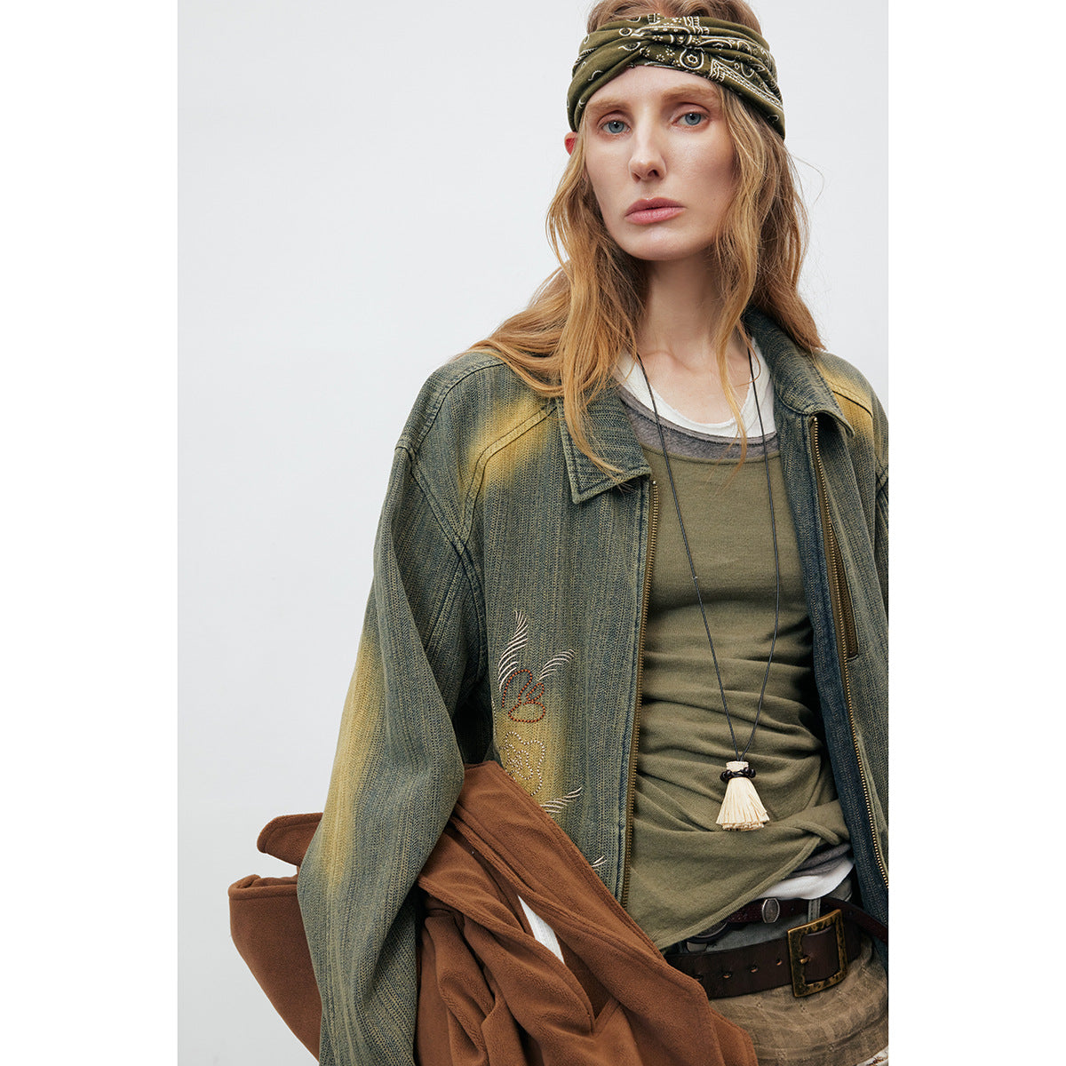 Close view of an olive distressed jacket with embroidered motifs, beside a green top and pendant necklace.