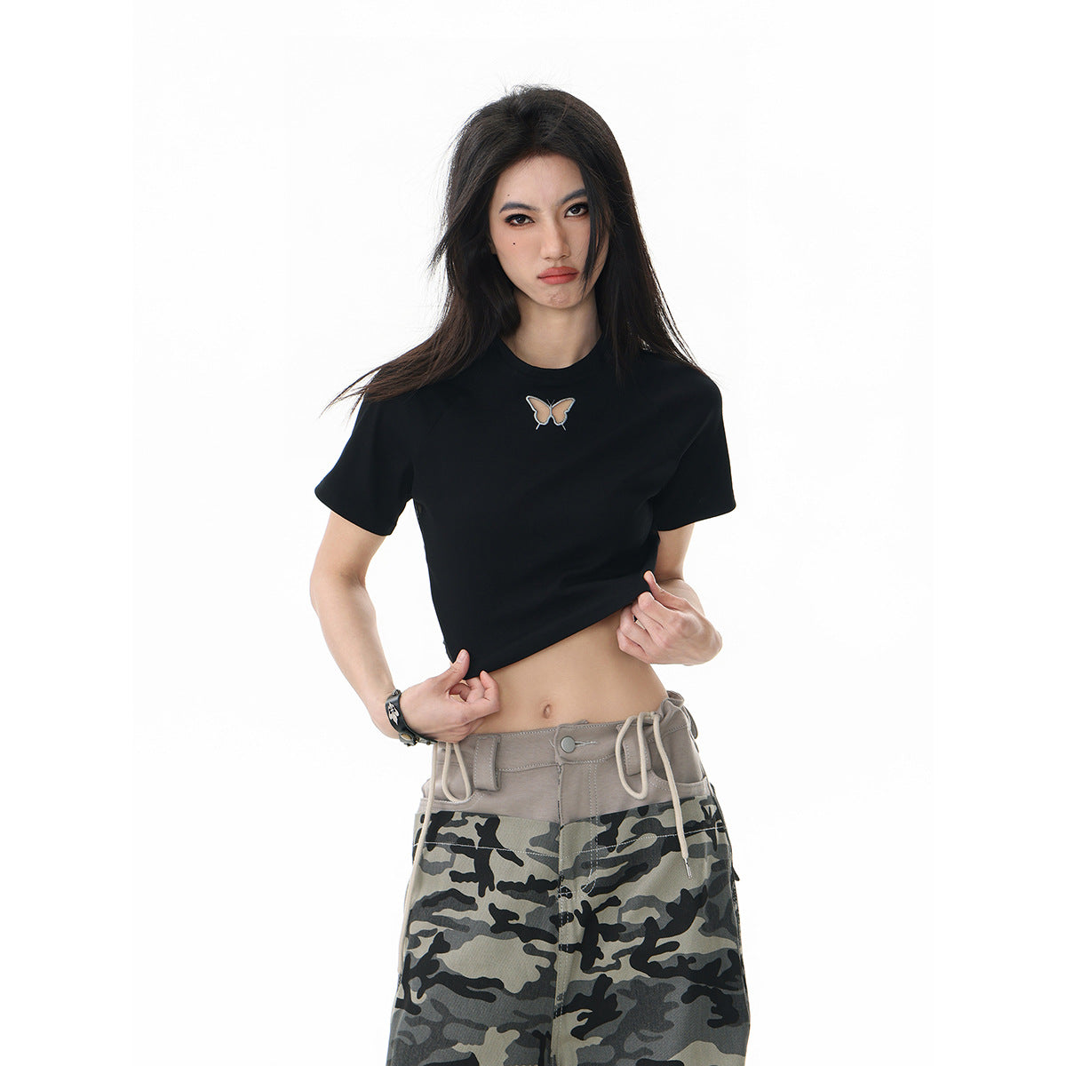 Model wearing a black cropped T-shirt with a small butterfly motif and loose gray camouflage pants.