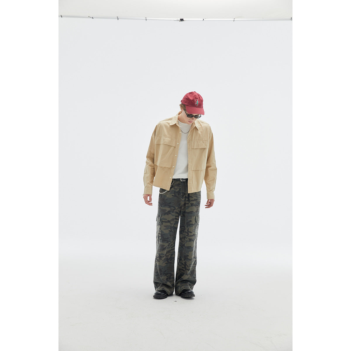 Model wearing a beige button-front overshirt, white top, and loose camouflage cargo pants with a red cap.