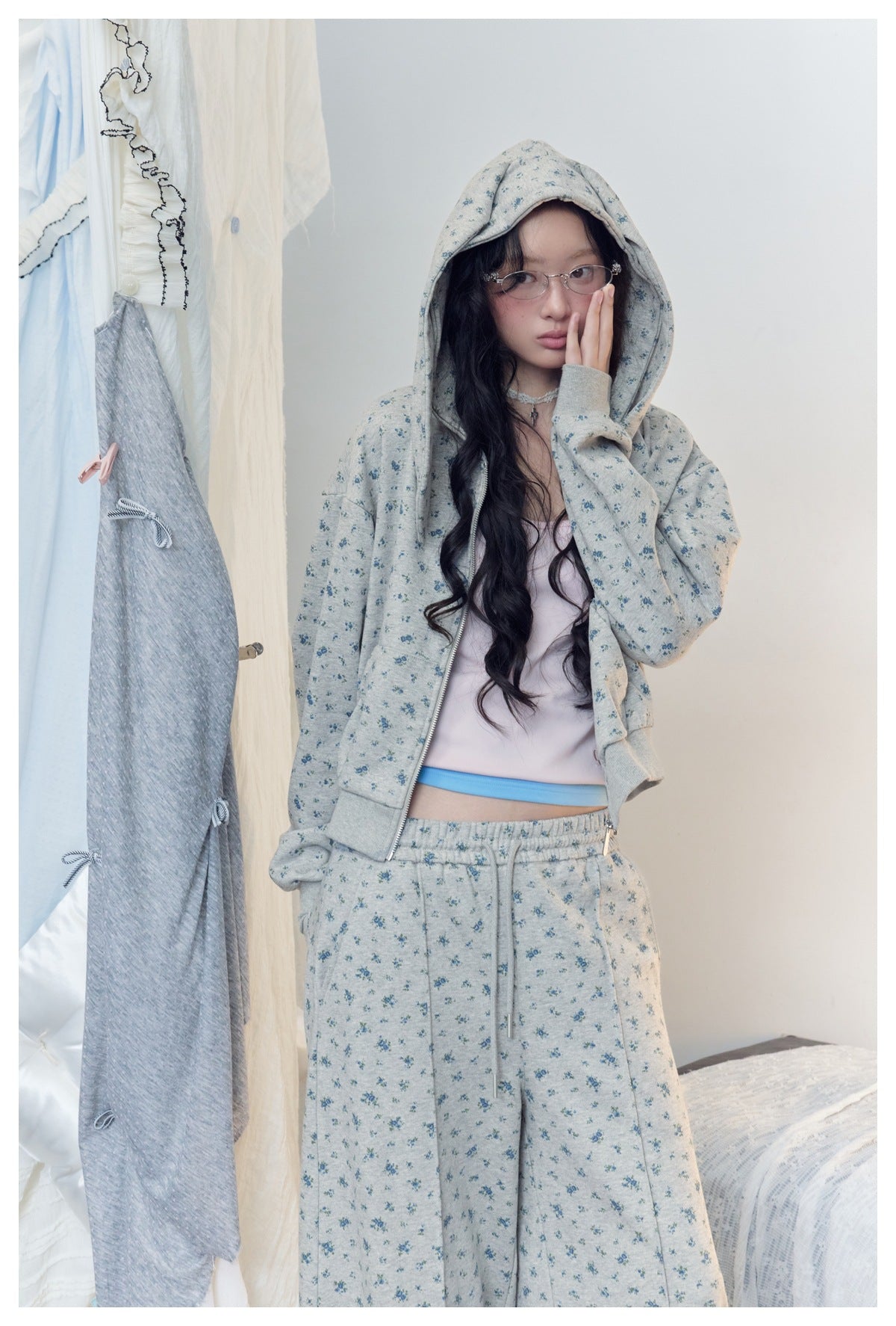 Model wearing a gray-blue floral hooded zip jacket and matching pants, with one hand raised beside her face.