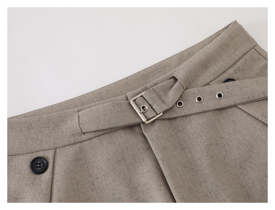Close-up of taupe trousers showing the belt buckle, metal eyelets, waistband, and front button.